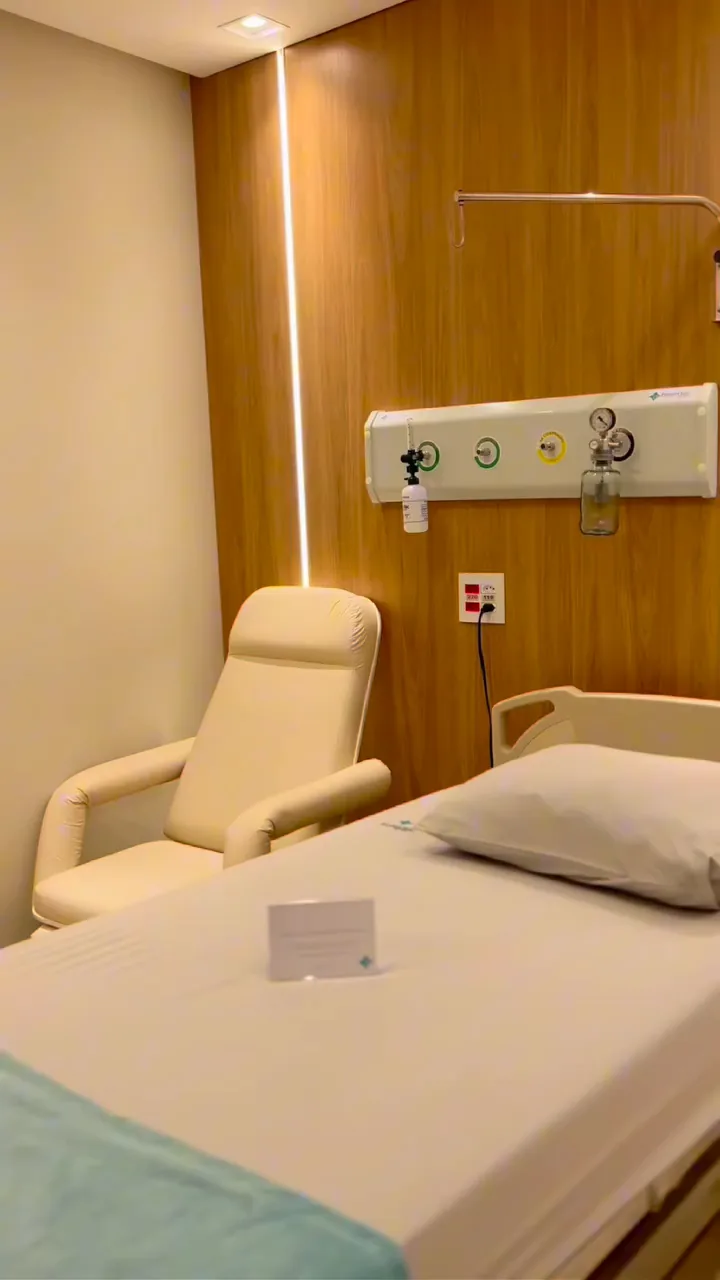 Quarto com leito, poltrona reclinável e painel de madeira com luz embutida.